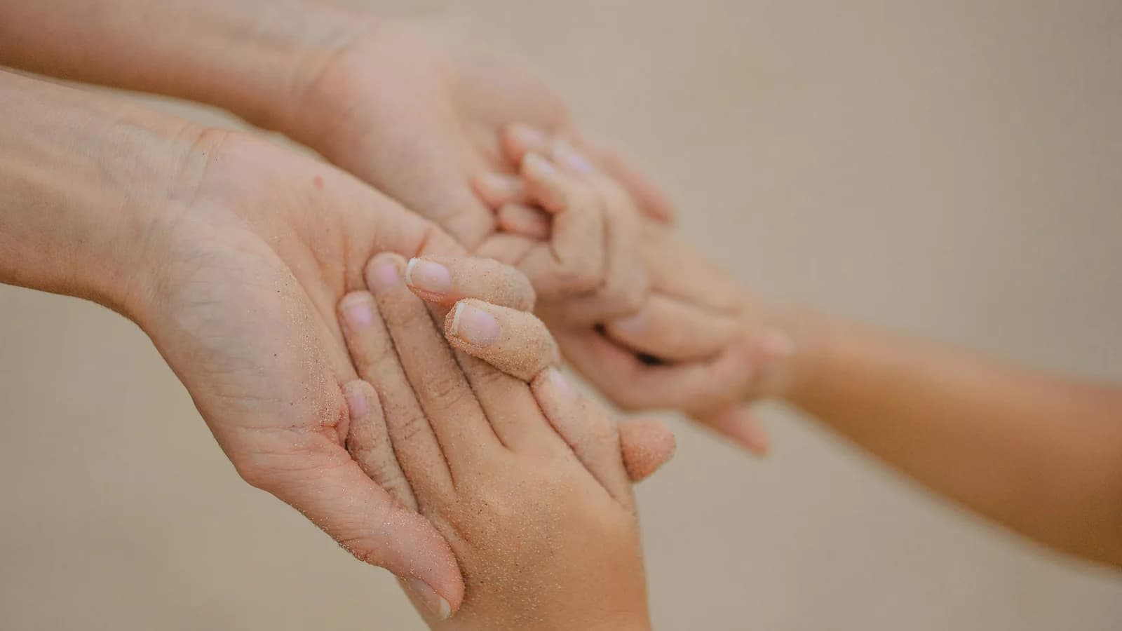 Deux mains d’adulte, couvertes de sable fin, tiennent délicatement les mains d’un enfant sur un fond clair et flou.