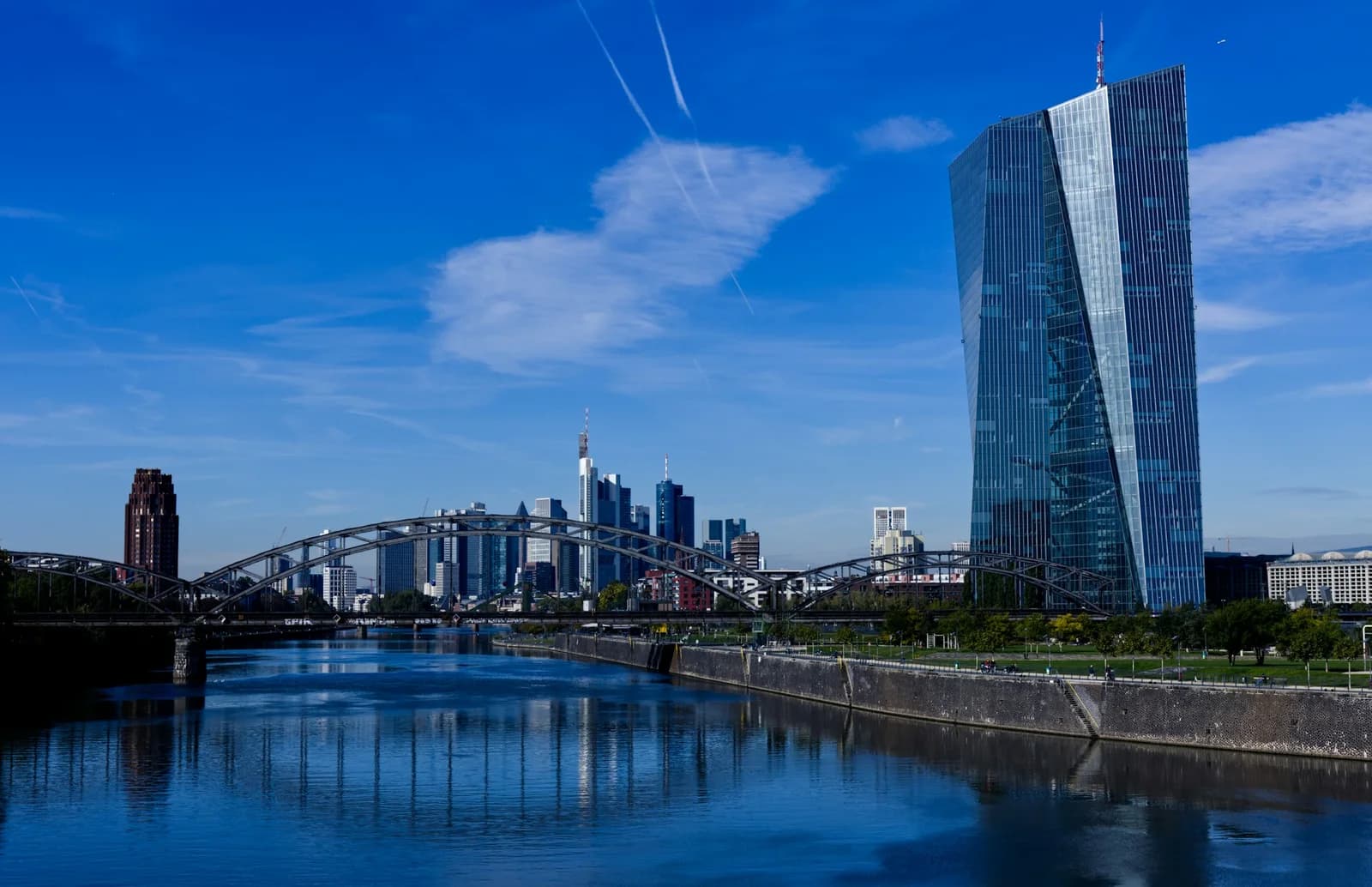 La tour de verre de la Banque centrale européenne au bord du Main, à Francfort, avec un pont métallique en arc et les gratte-ciel du quartier financier sous un ciel bleu.
