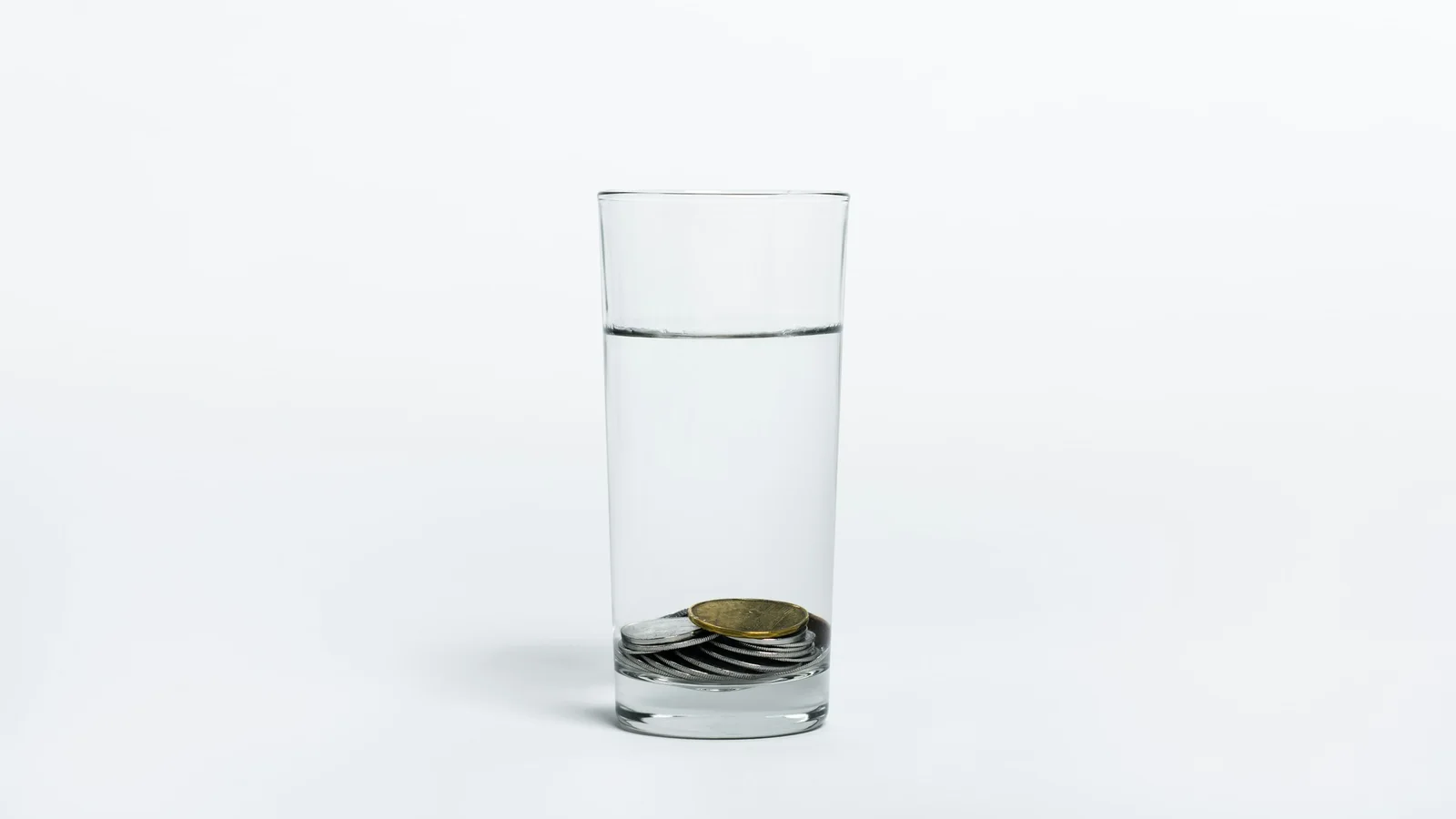 Un grand verre d’eau posé sur un fond blanc, au fond duquel reposent quelques pièces de monnaie.