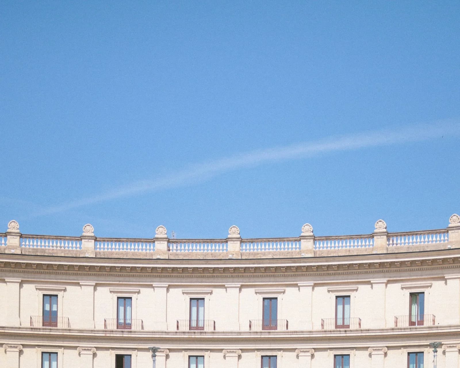 Façade classique incurvée, couronnée d’une balustrade de pierre claire, sous un vaste ciel bleu traversé d’une traînée blanche
