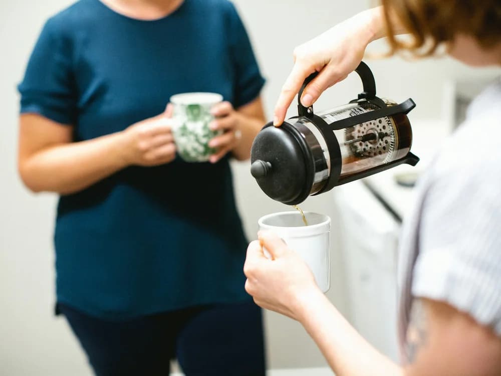 Pause café entre collègues : une main verse le café d’une cafetière à piston dans une tasse, une autre tient un mug.