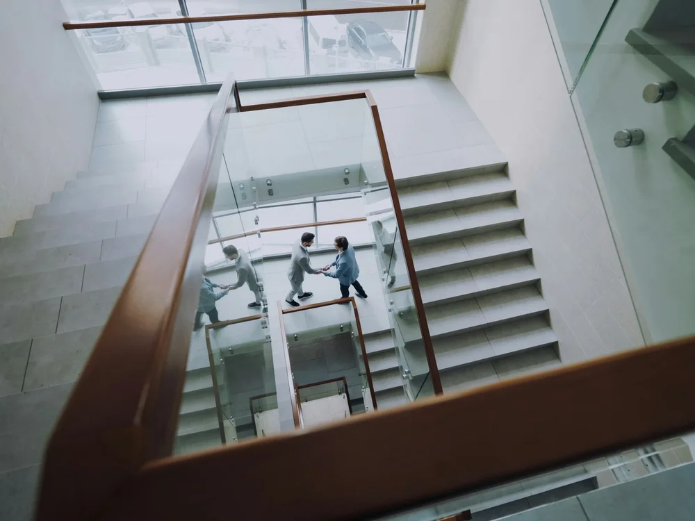 Vue plongeante sur une cage d’escalier moderne où deux collègues, tout petits dans le cadre, se serrent la main sur un palier vitré.