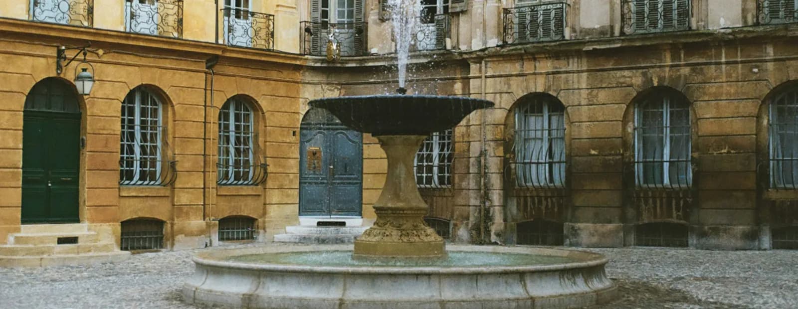 La fontaine de la place d’Albertas à Aix-en-Provence, entourée de façades classiques aux volets gris et aux balcons en ferronnerie.