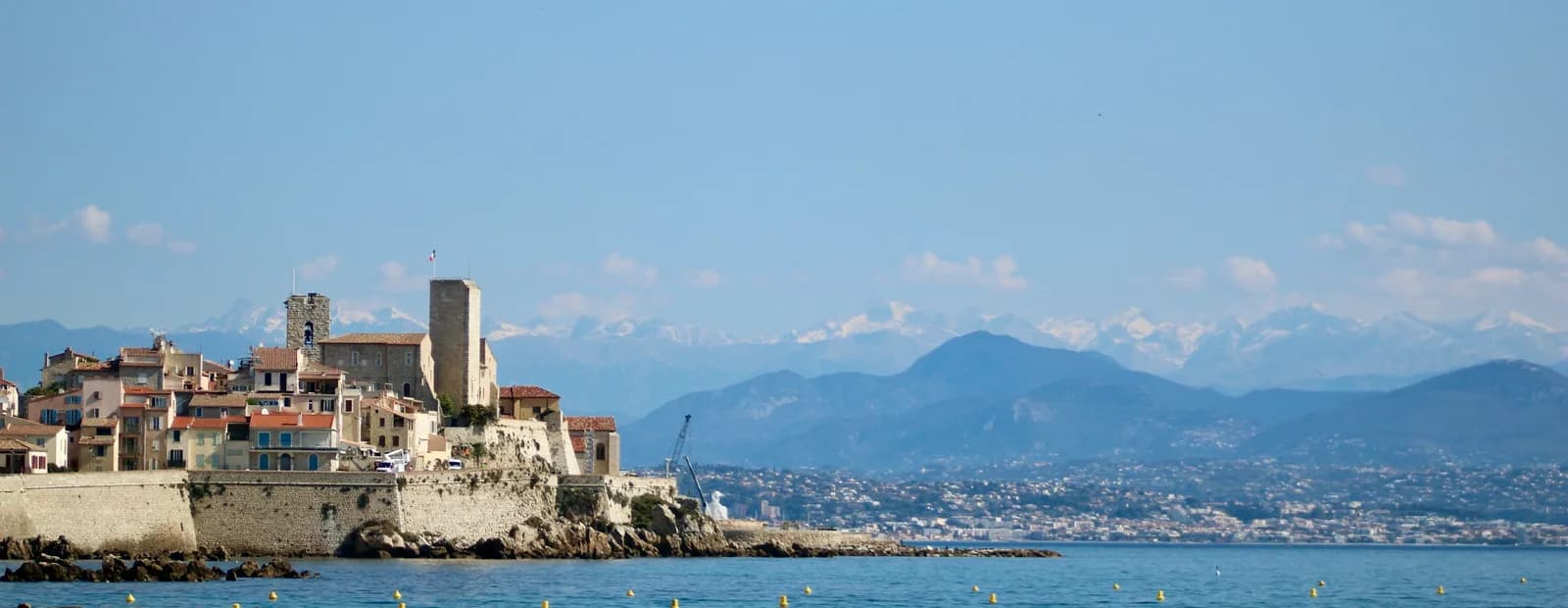 La vieille ville fortifiée d’Antibes et ses remparts au bord de la mer, avec les Alpes enneigées au loin sous un ciel bleu.