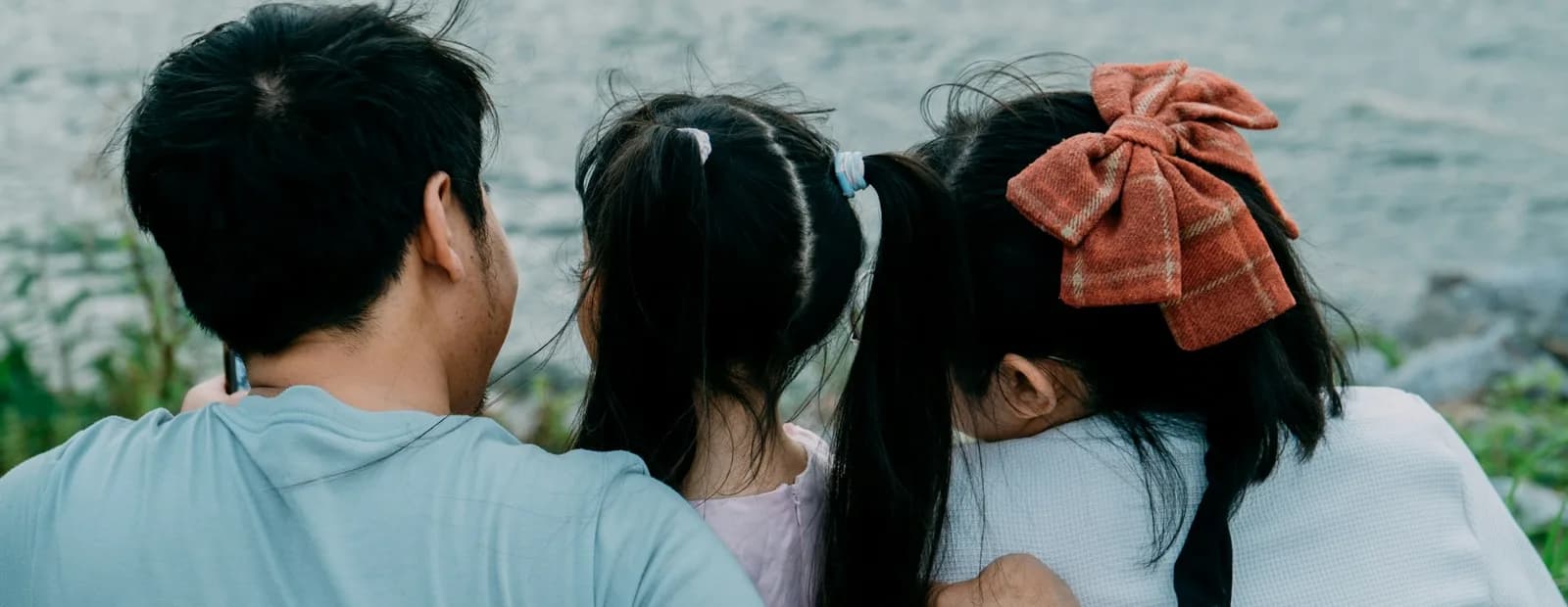 Un père et ses deux filles assis côte à côte, vus de dos, face à une étendue d’eau.