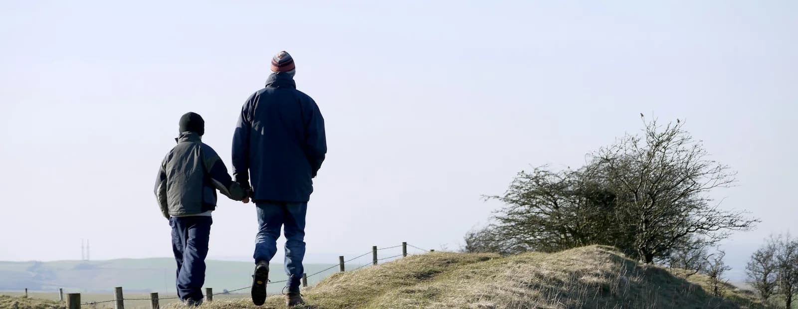 Un adulte et un enfant marchent main dans la main, de dos, sur un chemin herbeux en crête sous un ciel pâle.