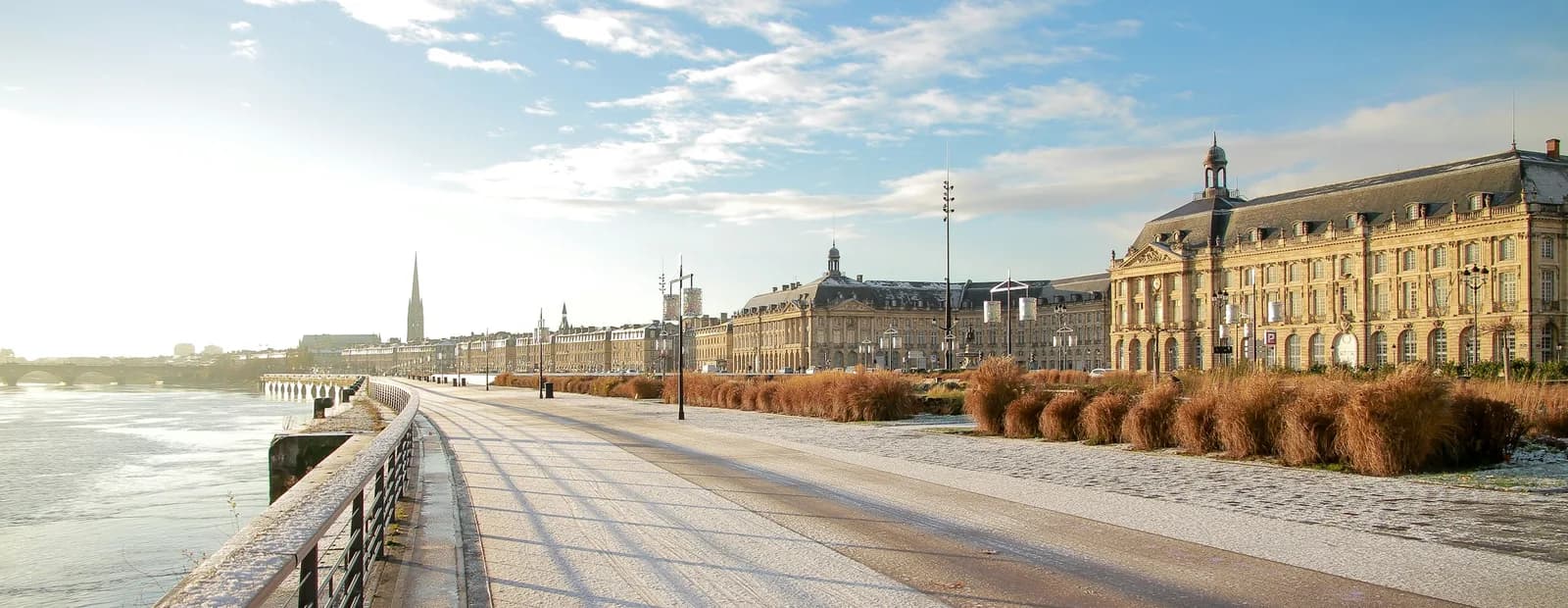 Les quais de Bordeaux sous une fine neige, le long de la Garonne, avec les façades classiques de la place de la Bourse au soleil.