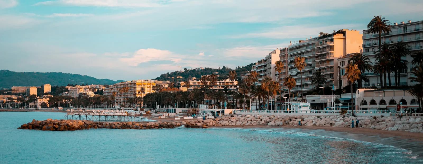 Le front de mer de Cannes au lever du jour : plage, digue de rochers, palmiers et immeubles clairs sous un ciel turquoise voilé.