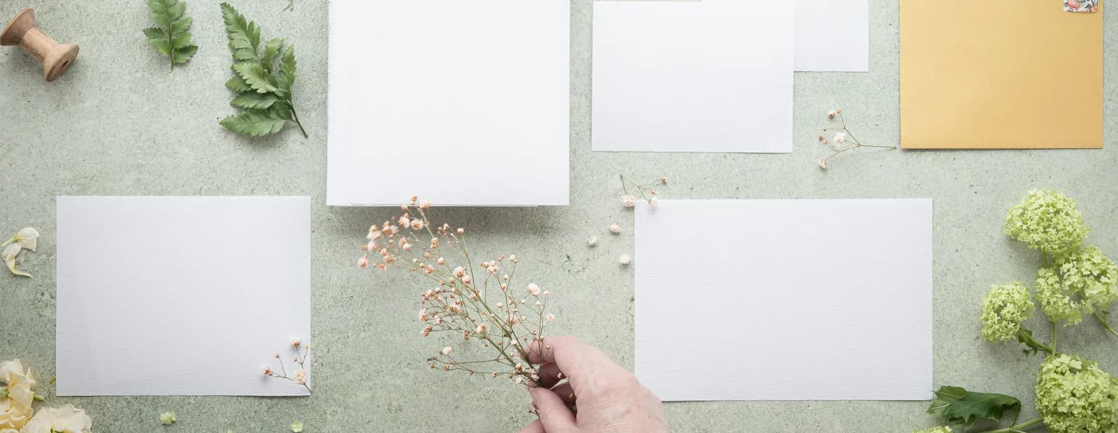 Cartes blanches, enveloppe dorée et brins de fleurs séchées disposés sur une table vert d’eau, une main ajustant la composition.