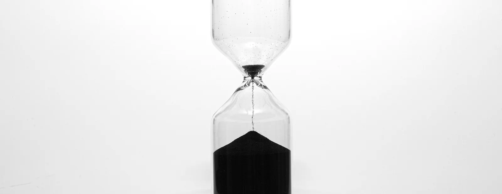 Sablier de verre sur fond blanc, un mince filet de sable noir s’écoulant vers le tas déjà formé.