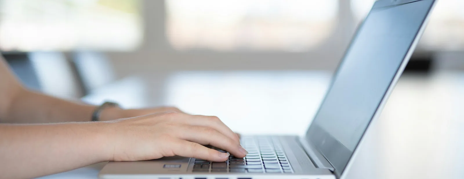 Mains posées sur le clavier d’un ordinateur portable vu de profil, écran éteint, sur une table blanche devant de grandes baies lumineuses floues.