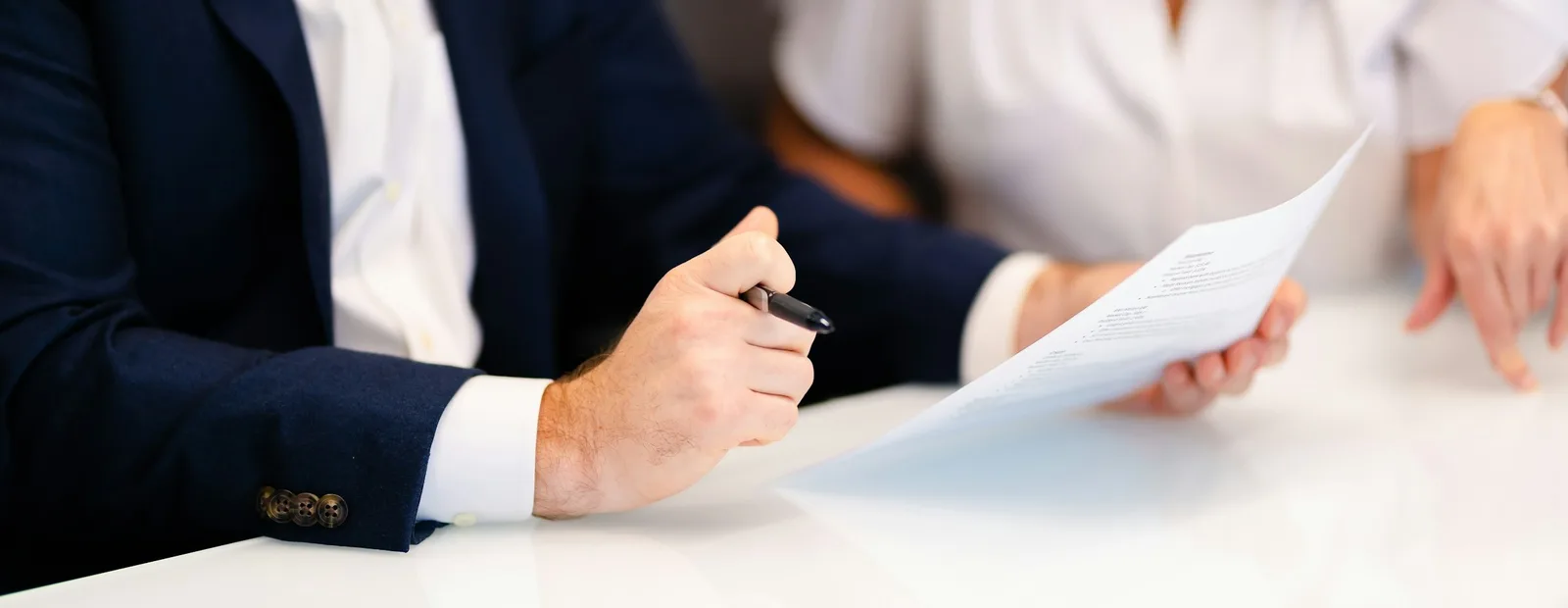 Deux personnes assises à une table blanche relisent un document : l’une, en veste bleu marine, tient un stylo, l’autre tient la feuille.