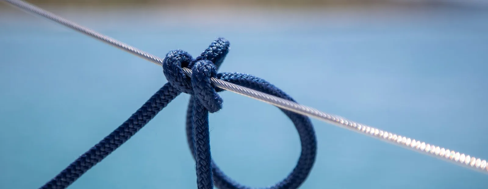 Nœud de cordage bleu marine serré sur un câble d’acier, devant une mer turquoise et une côte floues.