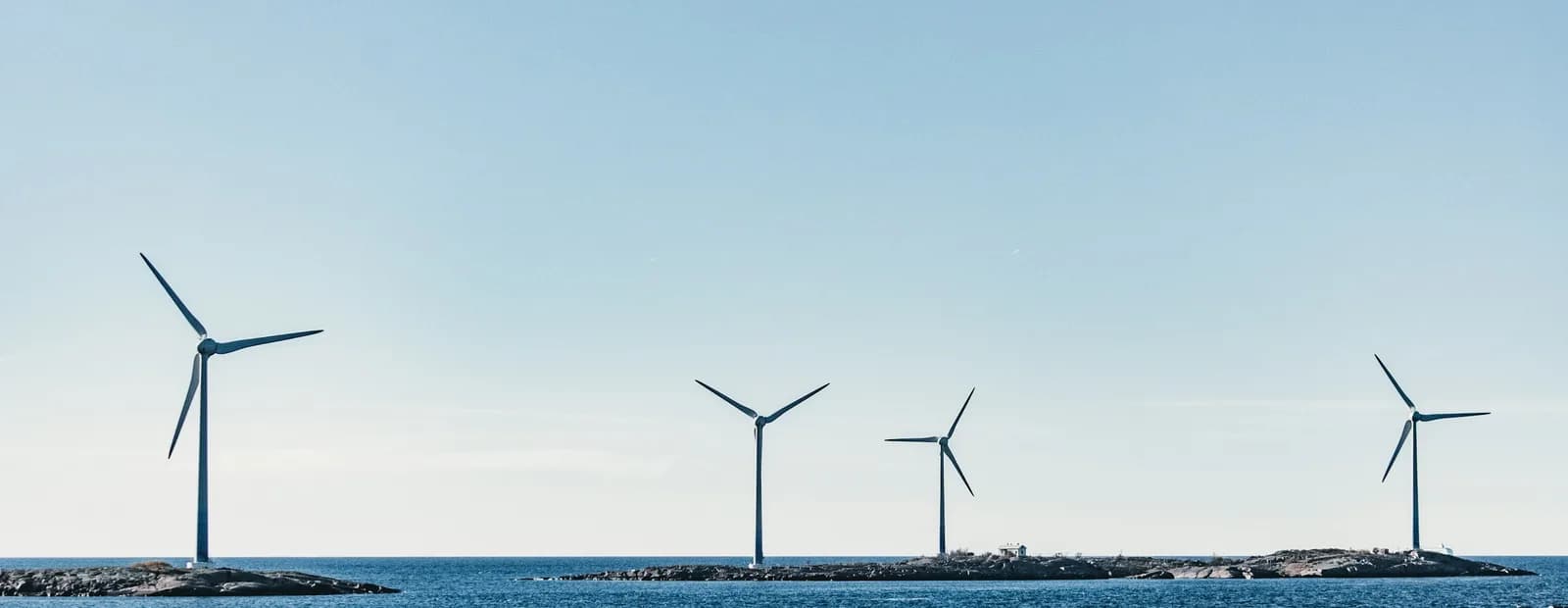 Quatre éoliennes plantées sur des îlots rocheux au ras d’une mer bleue, sous un ciel clair et dégagé