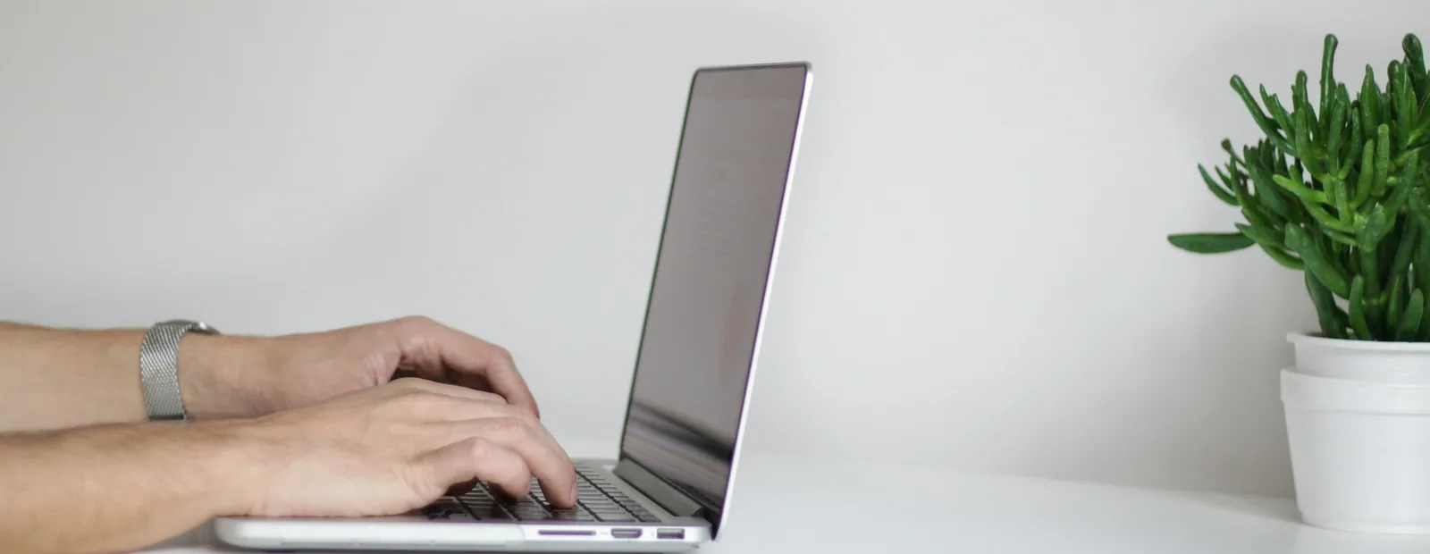 Des mains tapent sur un ordinateur portable posé sur un bureau blanc, à côté d’une plante verte en pot.