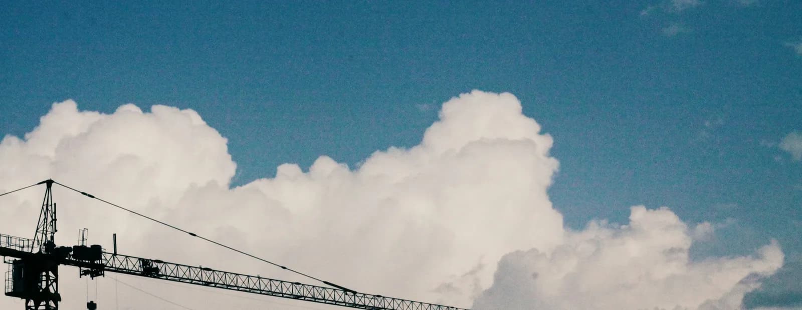 Silhouette d’une grue de chantier dont la flèche se détache sur de gros nuages blancs et un ciel bleu profond