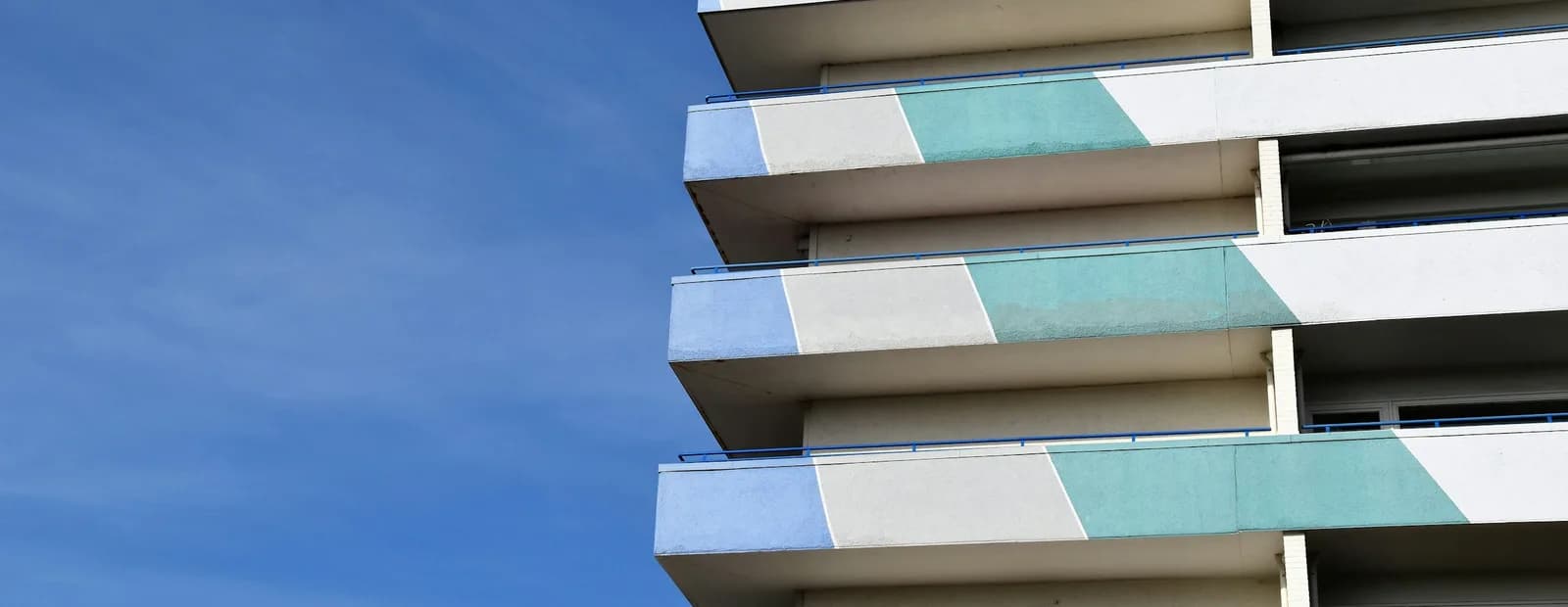 Angle d’un immeuble d’habitation récent aux balcons superposés à bandes bleues, vertes et blanches, sous un ciel bleu