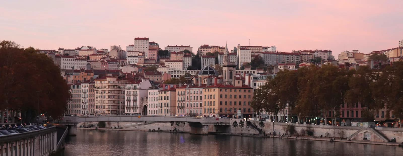 La Saône à Lyon au coucher du soleil, avec un pont, les façades des quais et les pentes de la Croix-Rousse sous un ciel rose.
