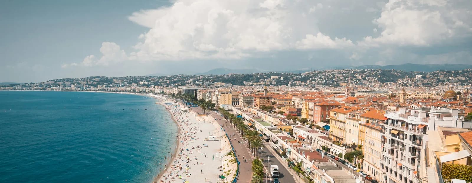 La baie des Anges et la promenade des Anglais à Nice vues d’en haut : mer bleue, longue plage de galets et façades claires sous un ciel nuageux.