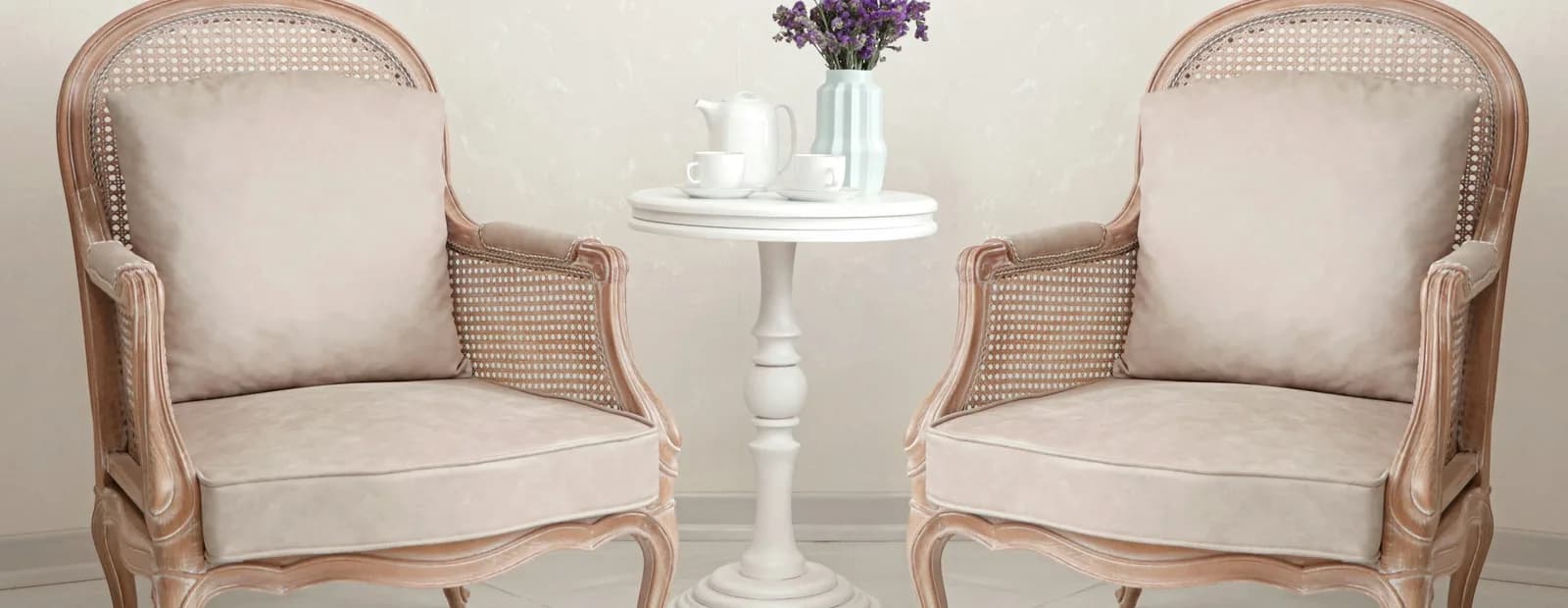 Deux fauteuils cannés face à face autour d’un guéridon blanc portant un service à thé et un bouquet de fleurs mauves, dans un salon clair.