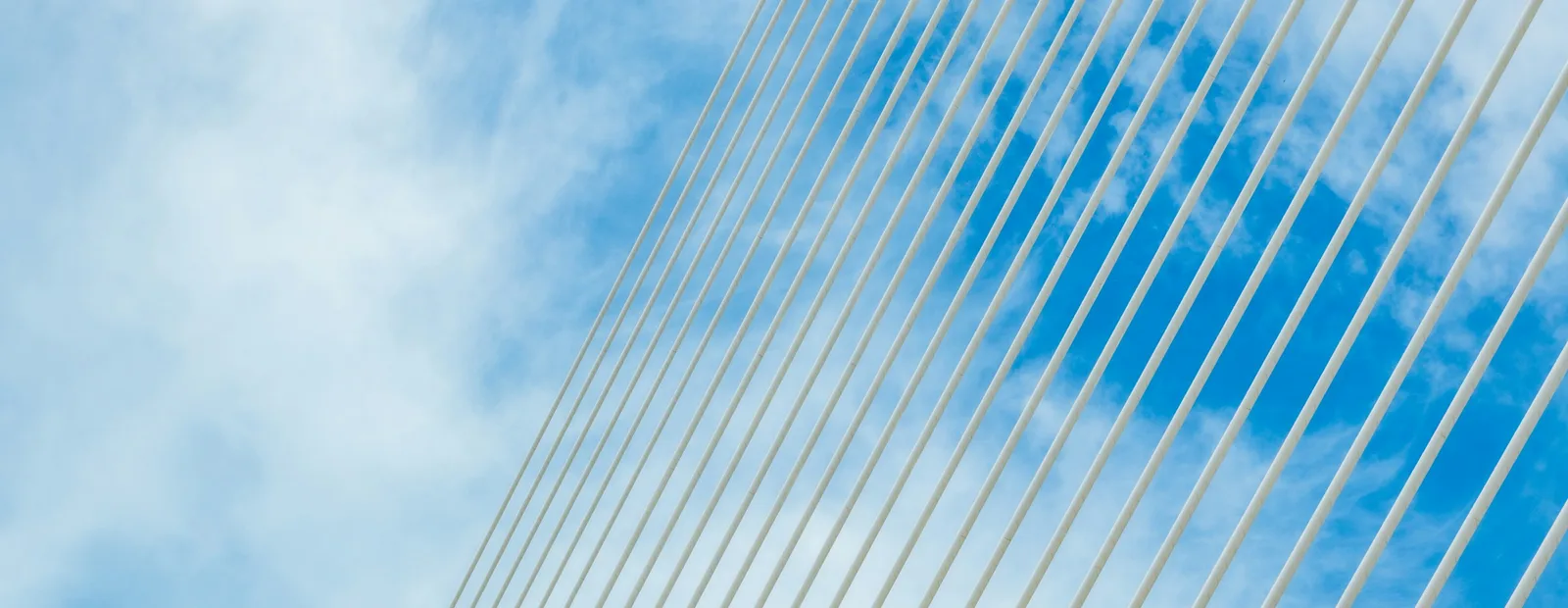 Faisceau de haubans blancs parallèles d’un pont suspendu, tendus en diagonale sur un ciel bleu traversé de nuages légers.