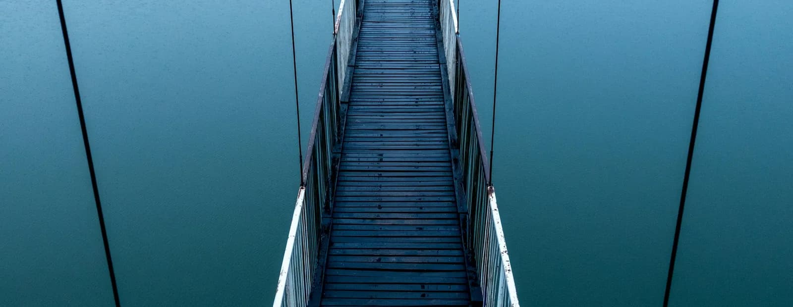 Passerelle suspendue en planches de bois, vide, qui file en perspective au-dessus d’une eau calme bleu-vert