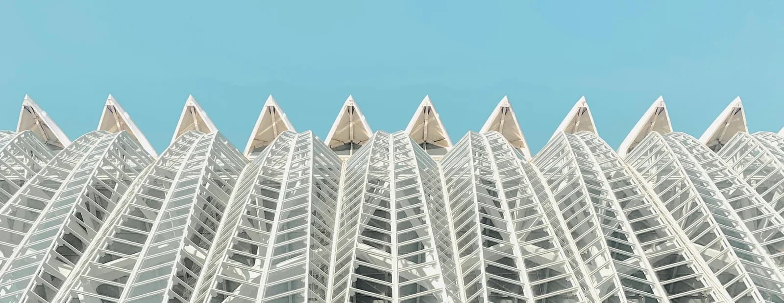 Façade blanche faite de modules vitrés identiques qui se répètent en rangs réguliers, coiffés de pointes, sous un ciel bleu pâle et uniforme.