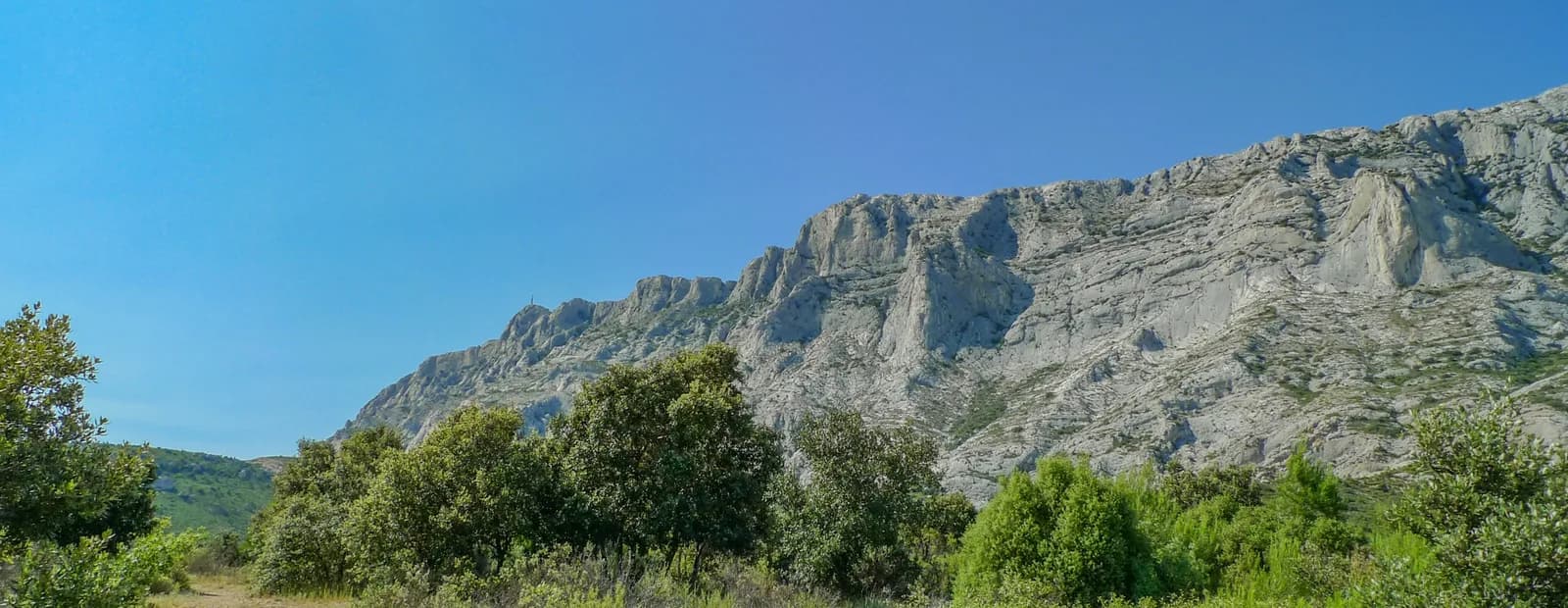 Les falaises calcaires de la montagne Sainte-Victoire sous un ciel bleu, au bout d’un chemin de terre bordé de garrigue.