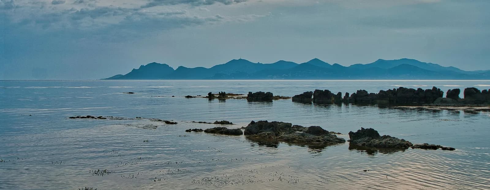 La silhouette bleutée du massif de l’Estérel vue depuis l’île Sainte-Marguerite, au-dessus d’une mer calme parsemée de rochers.
