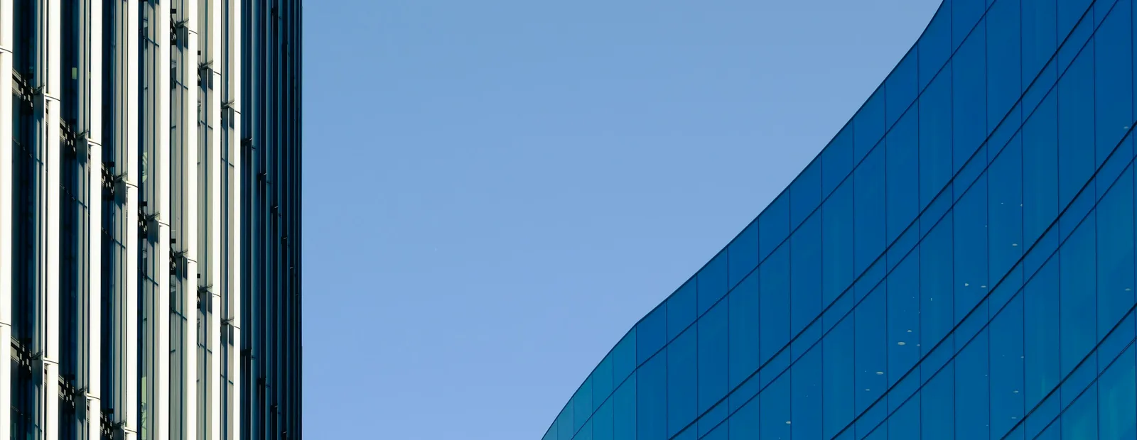 Deux immeubles de bureaux vitrés, l’un à lignes droites, l’autre à façade courbe bleue, sous un ciel bleu uni