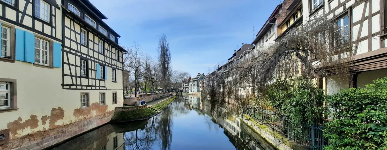 Un canal de la Petite France à Strasbourg bordé de maisons à colombages aux volets bleus, reflétées dans l’eau sous un ciel bleu.