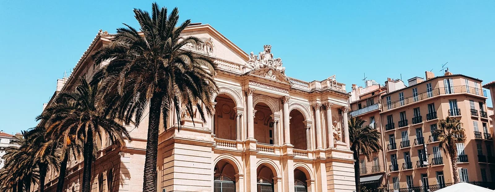 La façade à arcades de l’opéra de Toulon, bordée de grands palmiers, sous un ciel bleu uni ; immeubles clairs sur la droite.