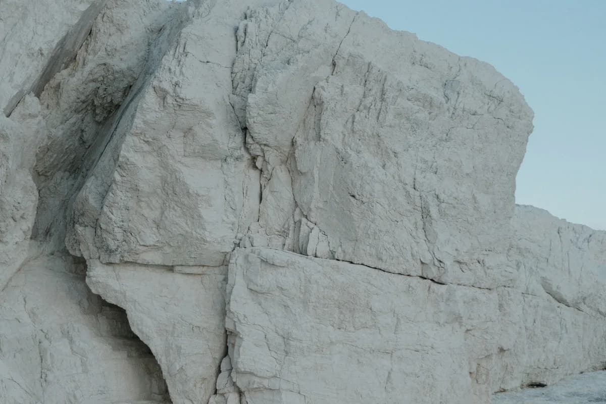 Gros bloc de roche calcaire blanche, brut et fissuré, posé sur un sol minéral clair sous un ciel bleu pâle sans nuage.