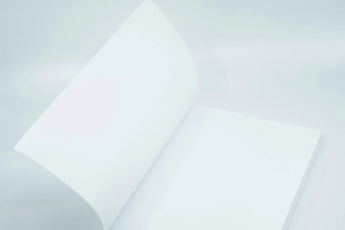 Livre ouvert aux pages entièrement blanches, une page en train de tourner, posé sur un fond de studio gris bleuté très clair.